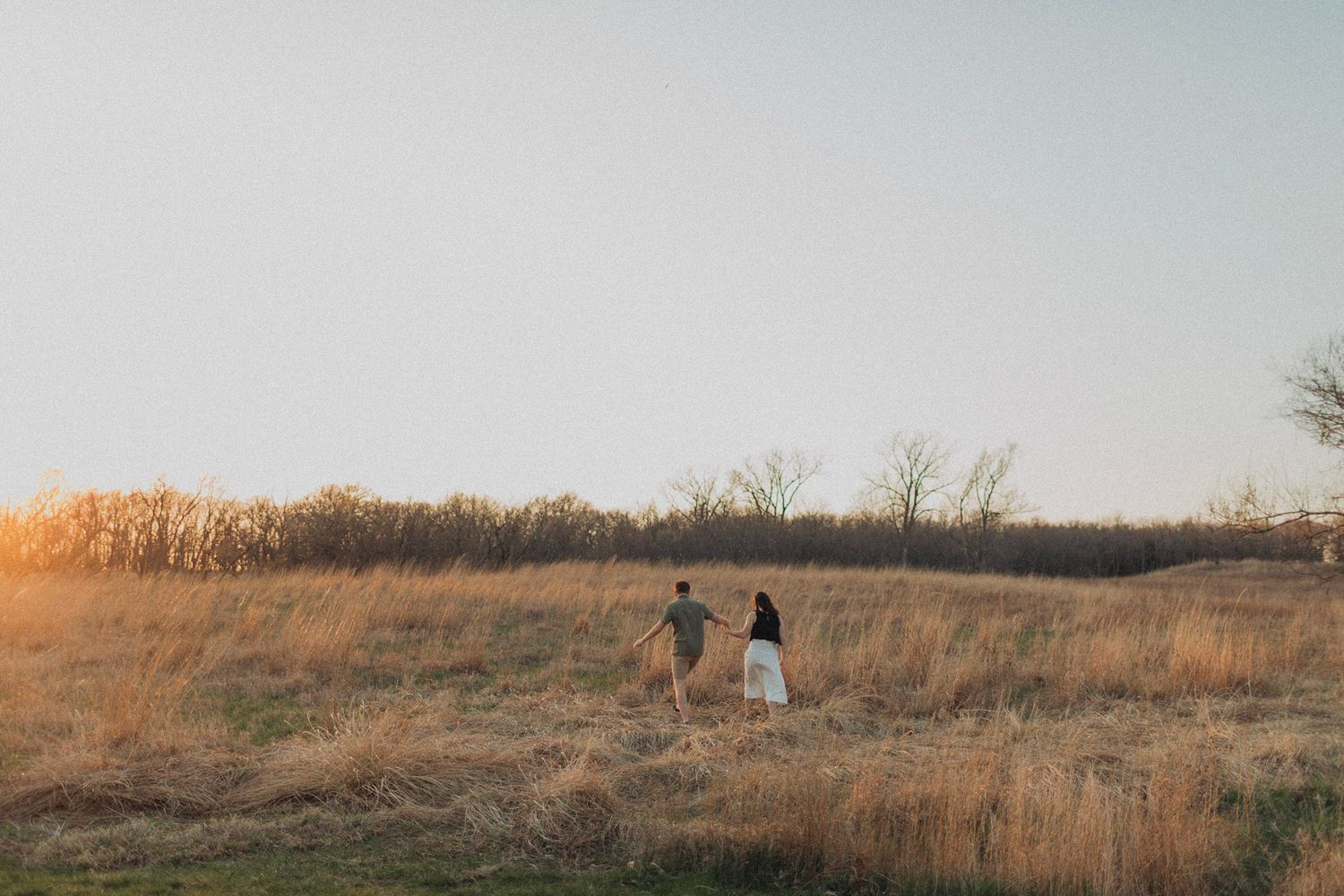 Couple holds hands walking through a golden prairie field at sunset in a wide open landscape in Montana Glacier National Park.
