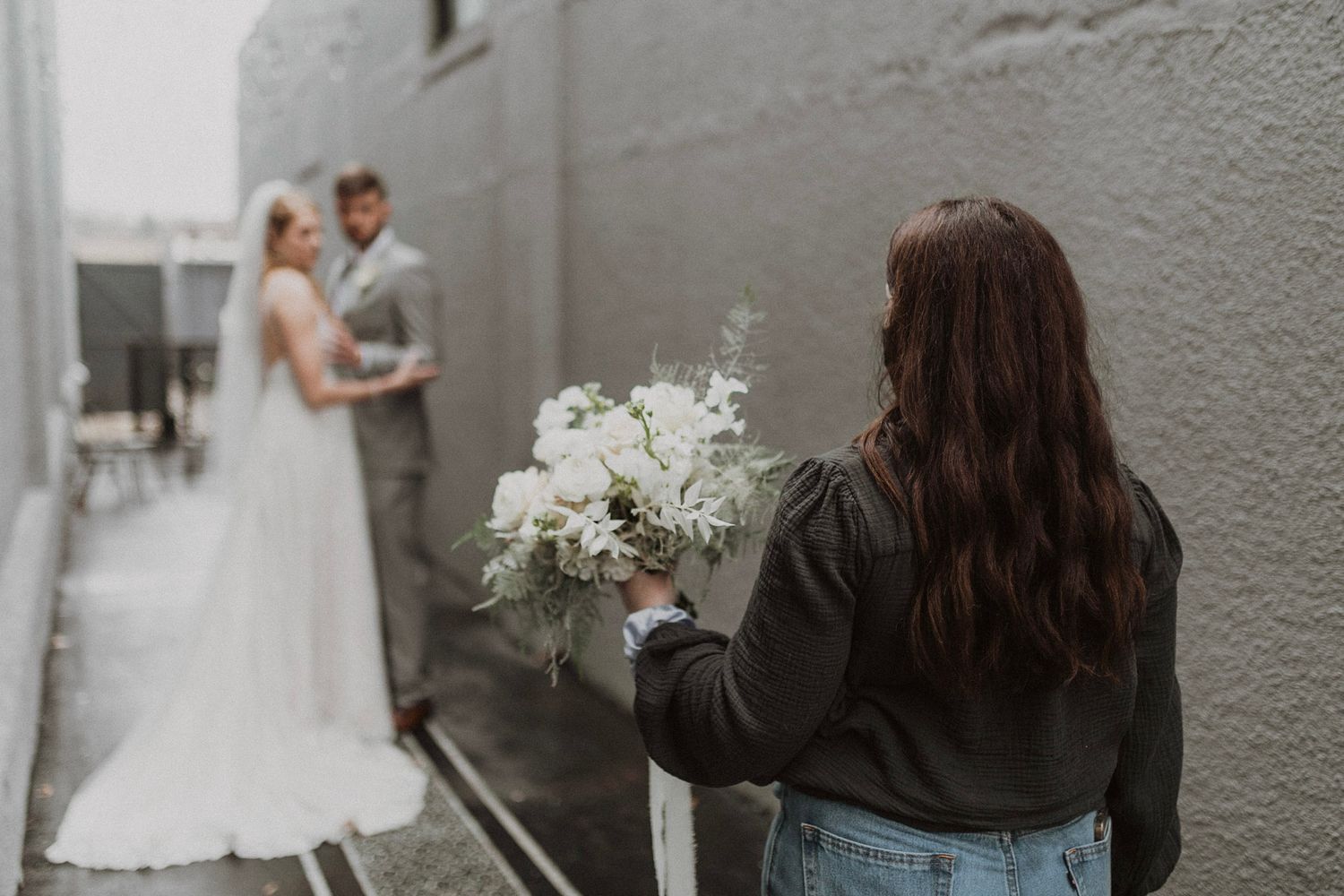 Wedding helper holds white floral bouquet while bride and groom pose against gray wall in alley in Montana Glacier National Park.