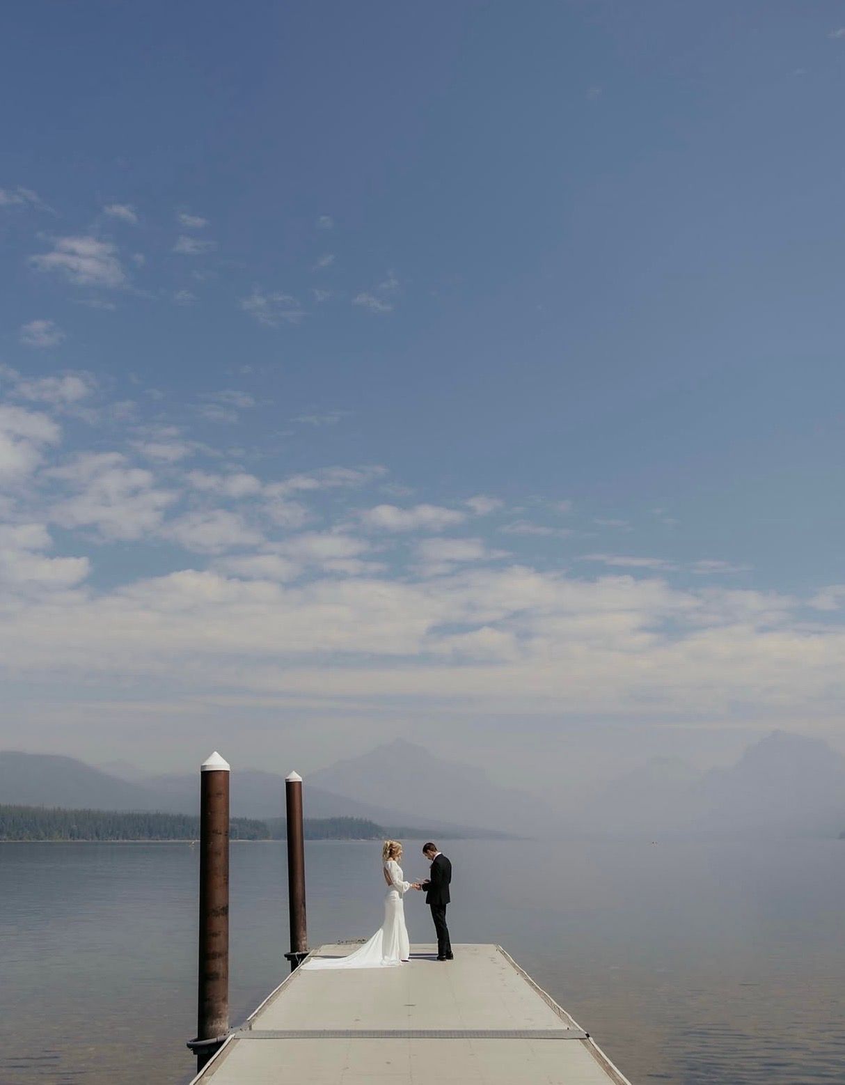 Bride and groom share an intimate moment on a serene lake dock under a vast blue sky in Montana Glacier National Park.