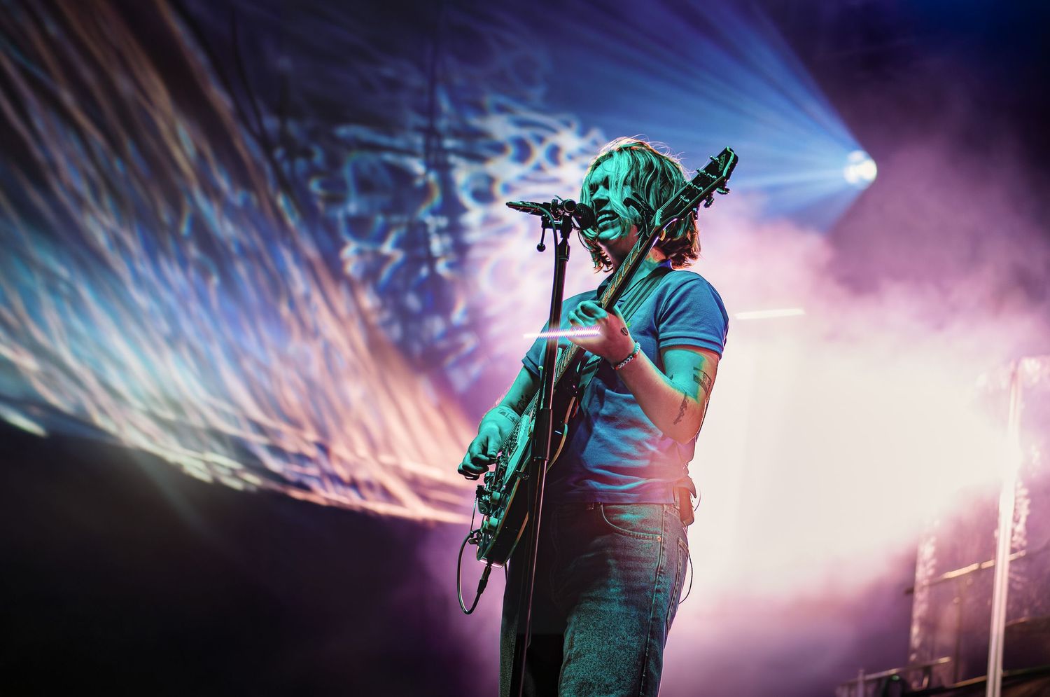 Guitarist singing into a microphone on stage with dramatic purple and blue lighting and projection backdrop.