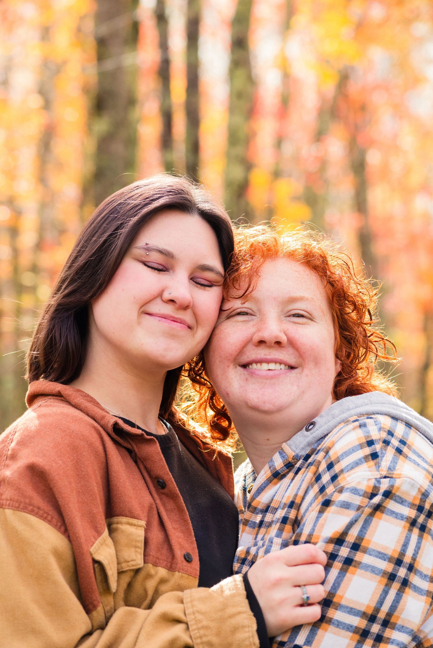 Two women smiling and embracing outdoors among vibrant autumn foliage in orange and yellow tones.