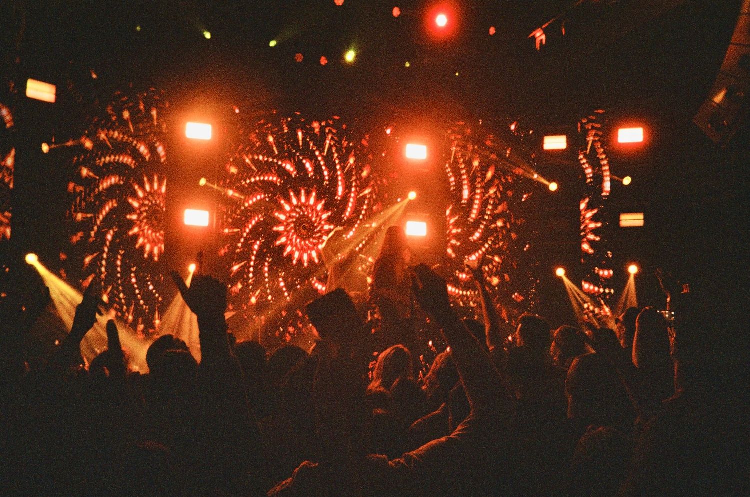 Energetic concert crowd with hands raised under spectacular red and orange spinning light displays.