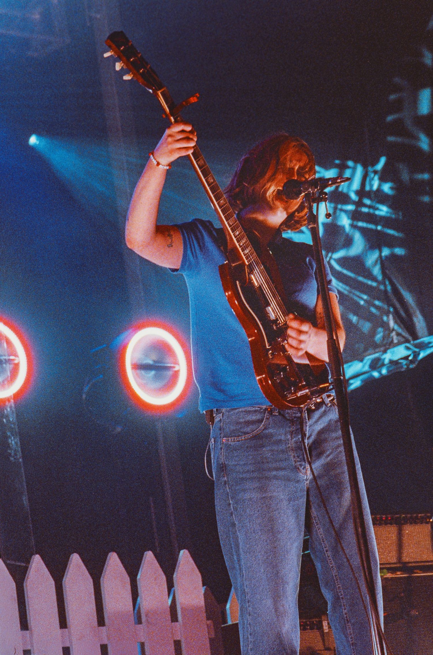 Guitarist lifting their guitar overhead on a colorful stage with neon circle lights and a white picket fence.
