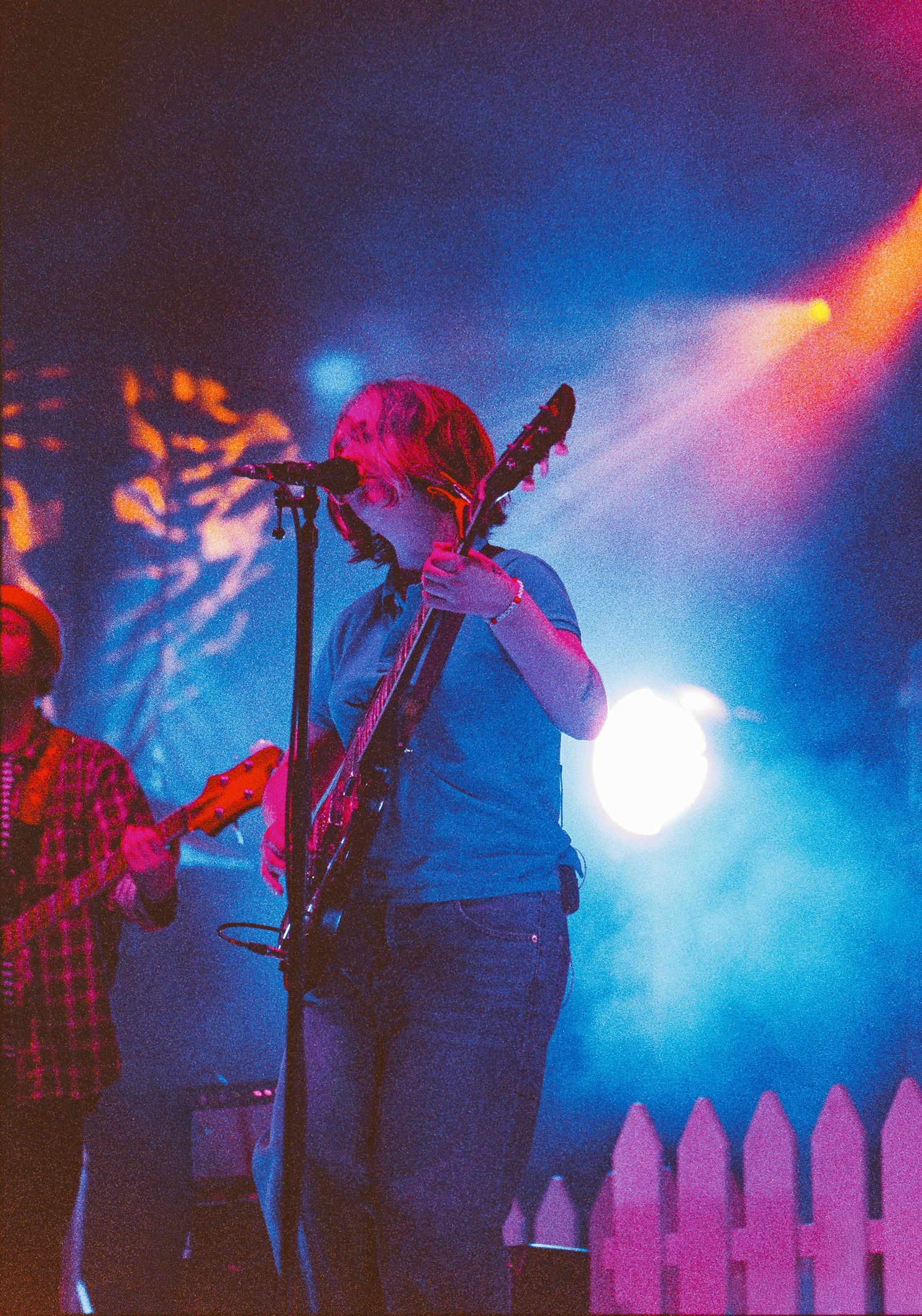 Guitarist performing on stage under blue and red lights with a white picket fence backdrop.