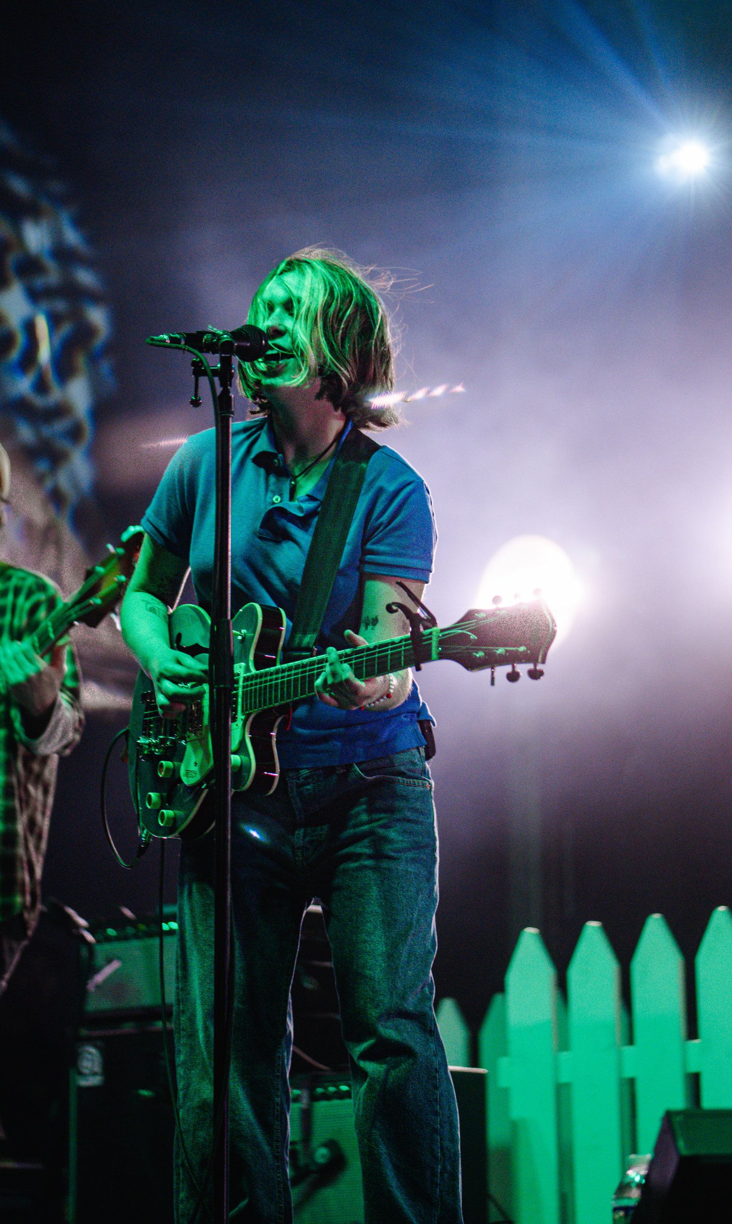 Guitarist singing into a microphone on a smoke-filled green-lit stage with a white picket fence backdrop.