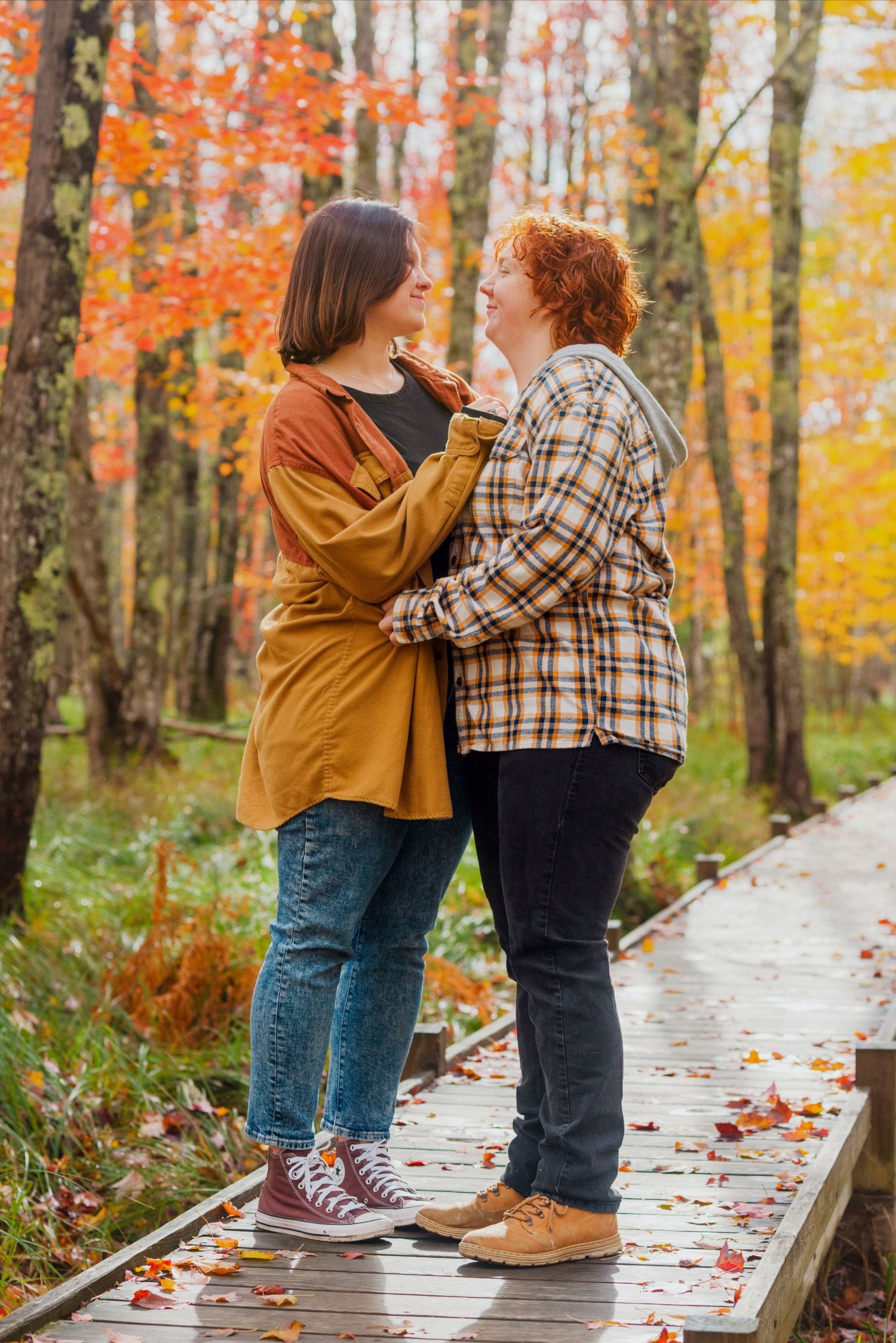 Two women embrace on a wooden boardwalk surrounded by vibrant autumn foliage in shades of red, orange, and yellow.