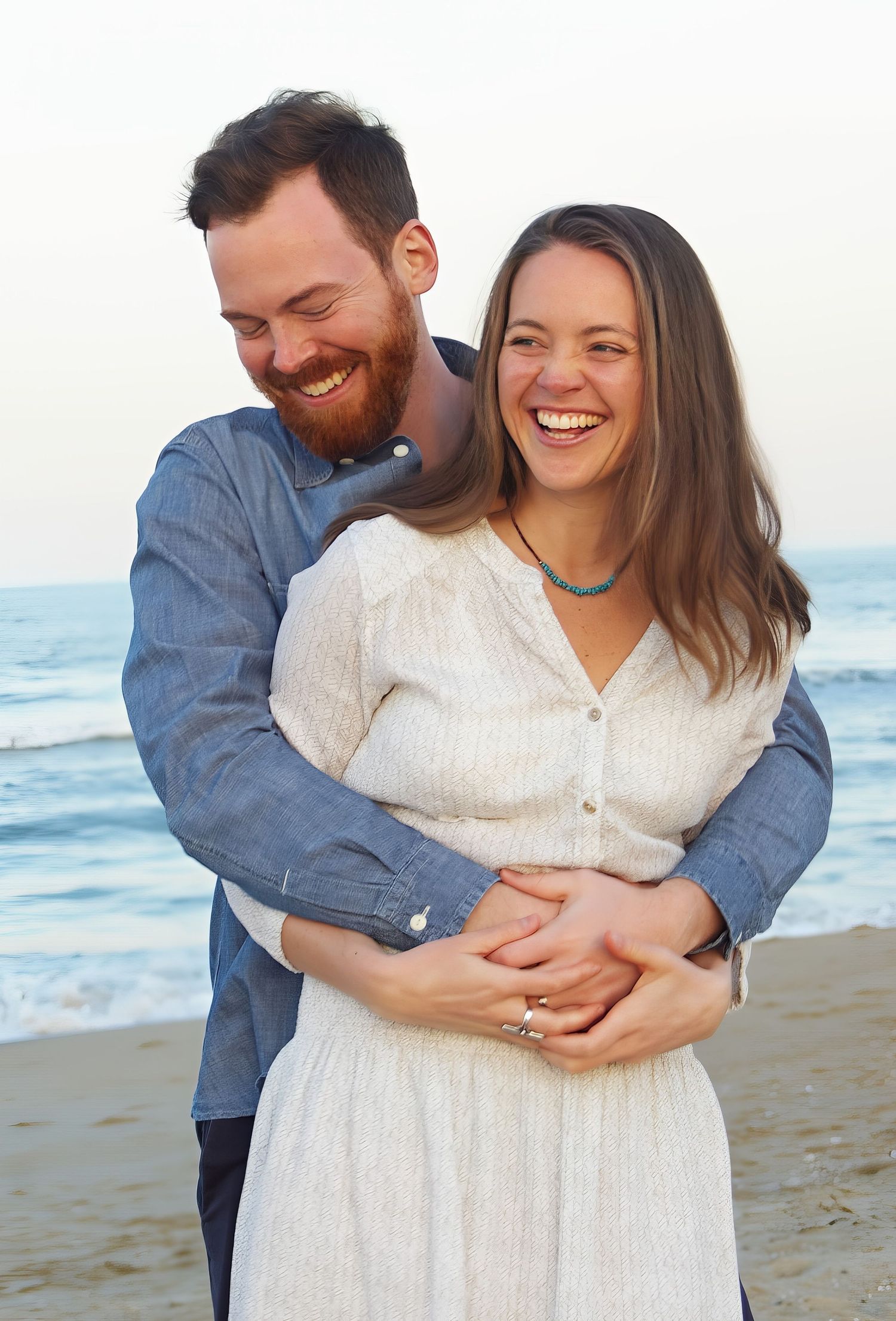 Happy couple embracing on a beach at sunset, woman in white dress with turquoise necklace, man in blue shirt smiling.
