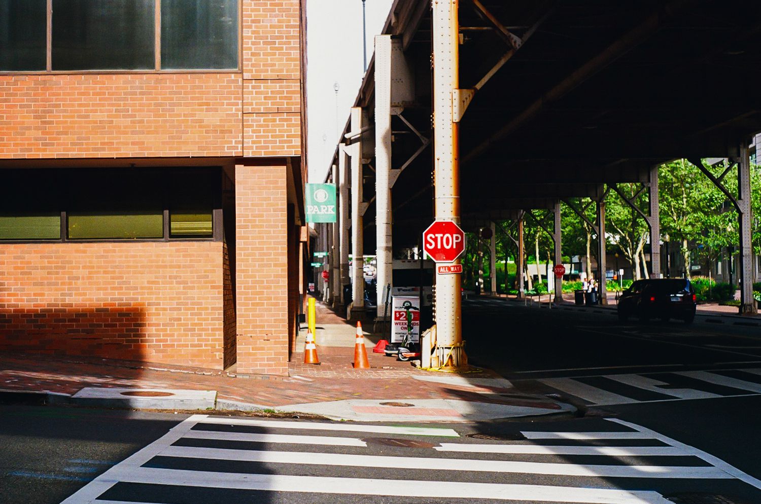 Urban street corner with a red stop sign, brick building, crosswalk, and elevated structure in a city setting.