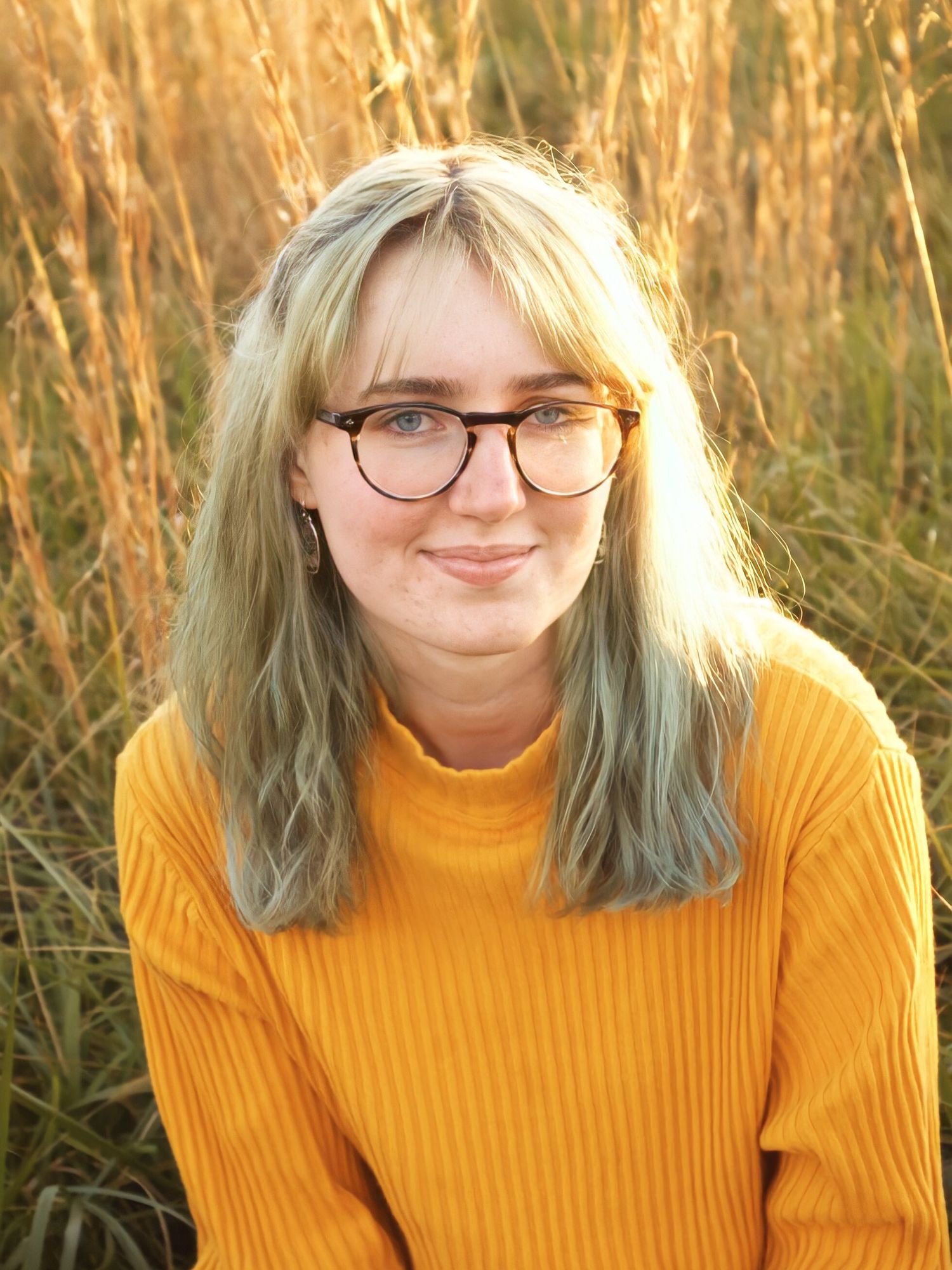 Young woman with blonde hair and glasses wearing a yellow turtleneck sweater, smiling outdoors in golden field.