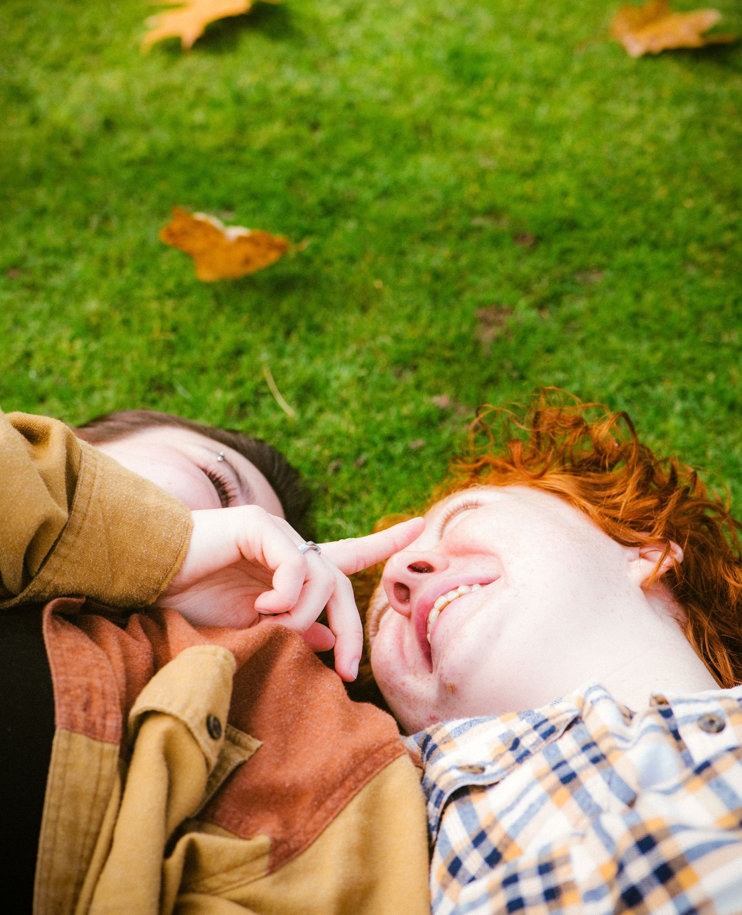Two laughing friends with red and dark hair lie on green grass in autumn, playfully touching each other's faces.