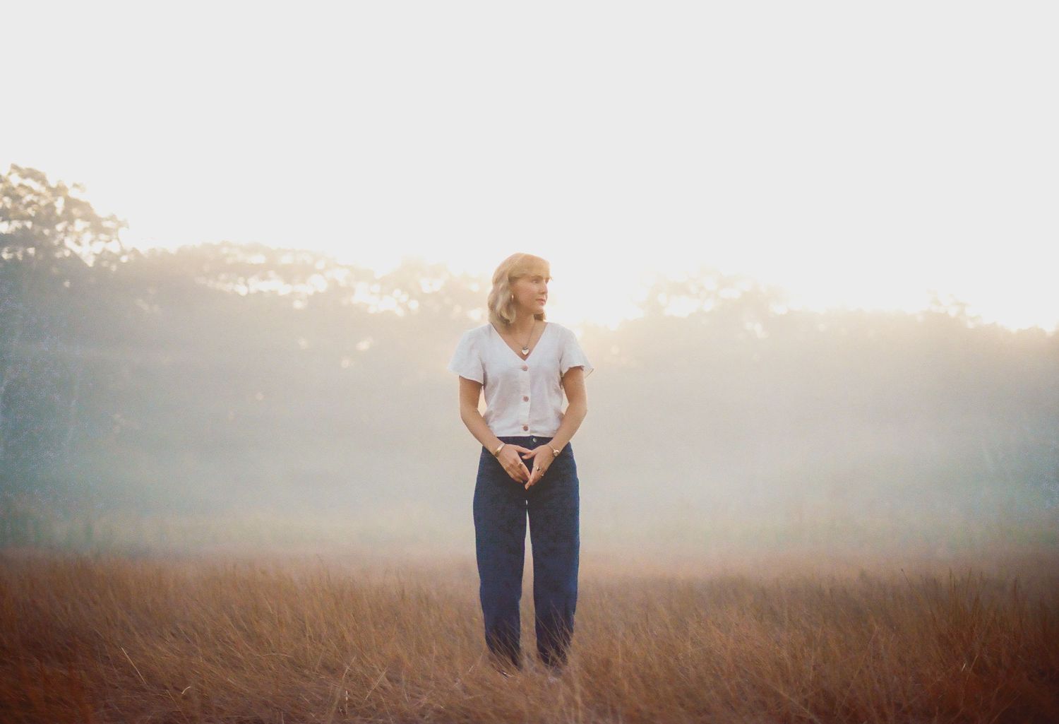 A woman in a white blouse and blue jeans stands peacefully in a sunlit misty meadow.