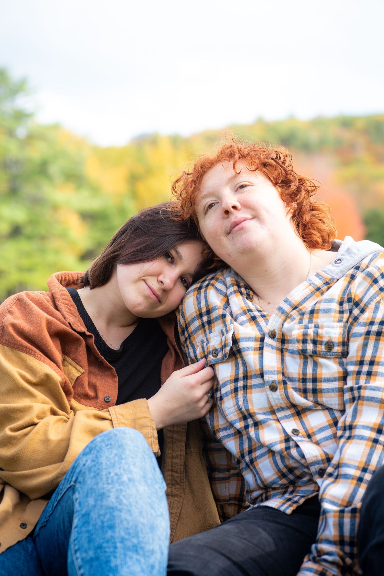 Two women with dark and curly red hair lean on each other outdoors in autumn, wearing plaid and yellow jackets.