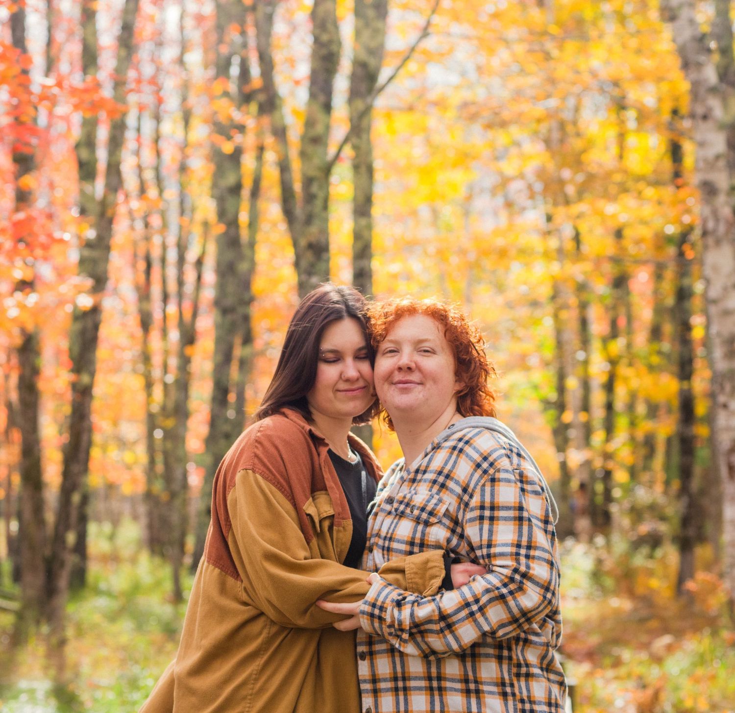 Two women embrace warmly in an autumn forest with vibrant orange and yellow foliage in the background.