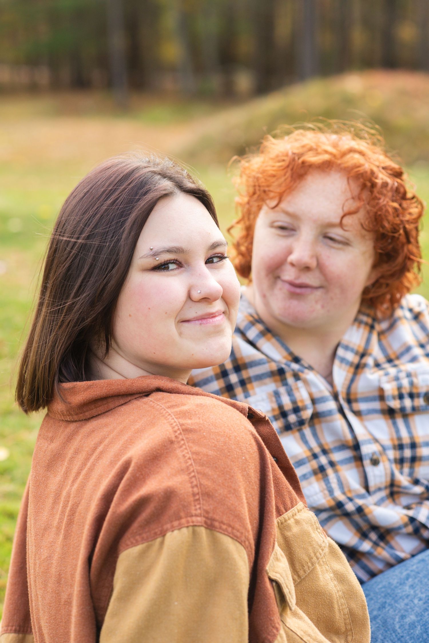 Two young women smiling outdoors in autumn, one with dark hair in orange jacket, other with curly red hair in plaid shirt.