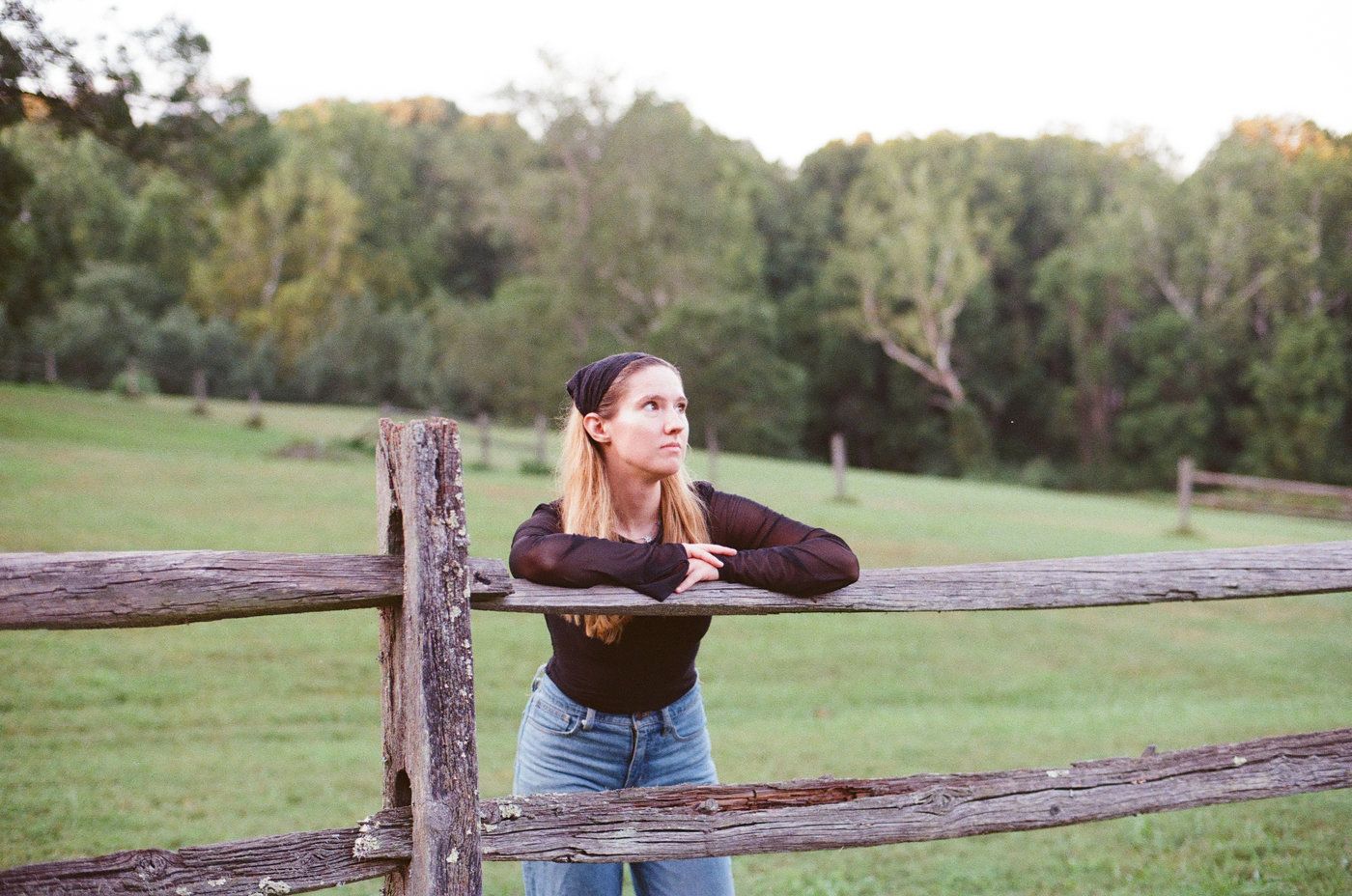 Young woman in jeans and black top leaning on rustic wooden fence in lush green countryside at sunset.