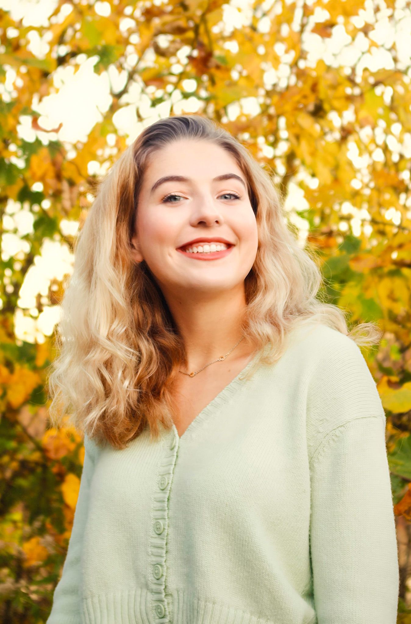 Smiling young woman with blonde wavy hair wearing a mint green cardigan outdoors with autumn foliage background.