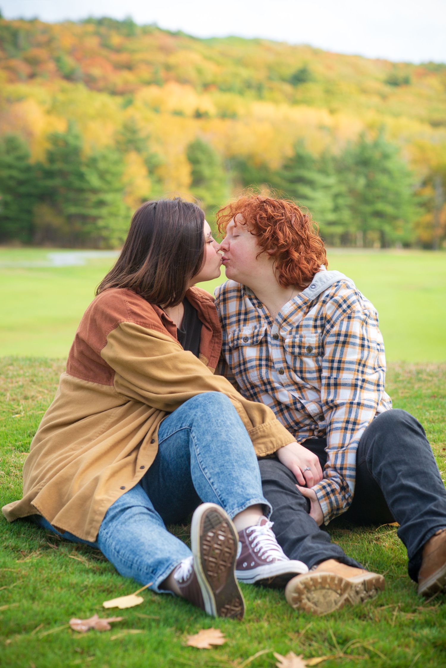 A couple shares a romantic kiss while sitting on green grass with colorful autumn foliage in the background.