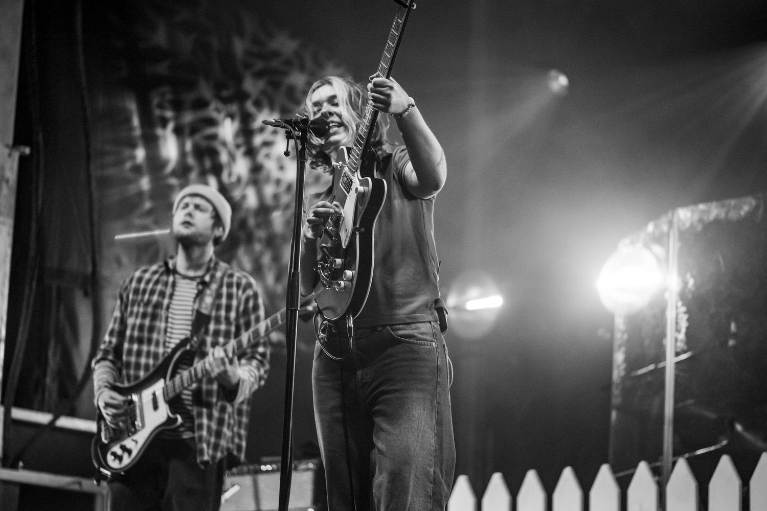 Two musicians perform on stage in black and white, one playing bass guitar and one playing electric guitar under dramatic stage lighting.