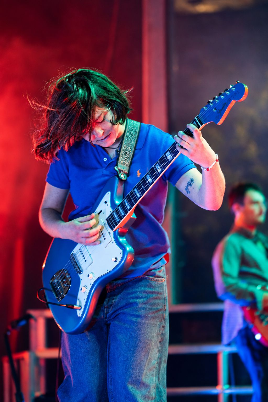 Female guitarist playing a blue electric guitar on stage under red and green concert lighting.
