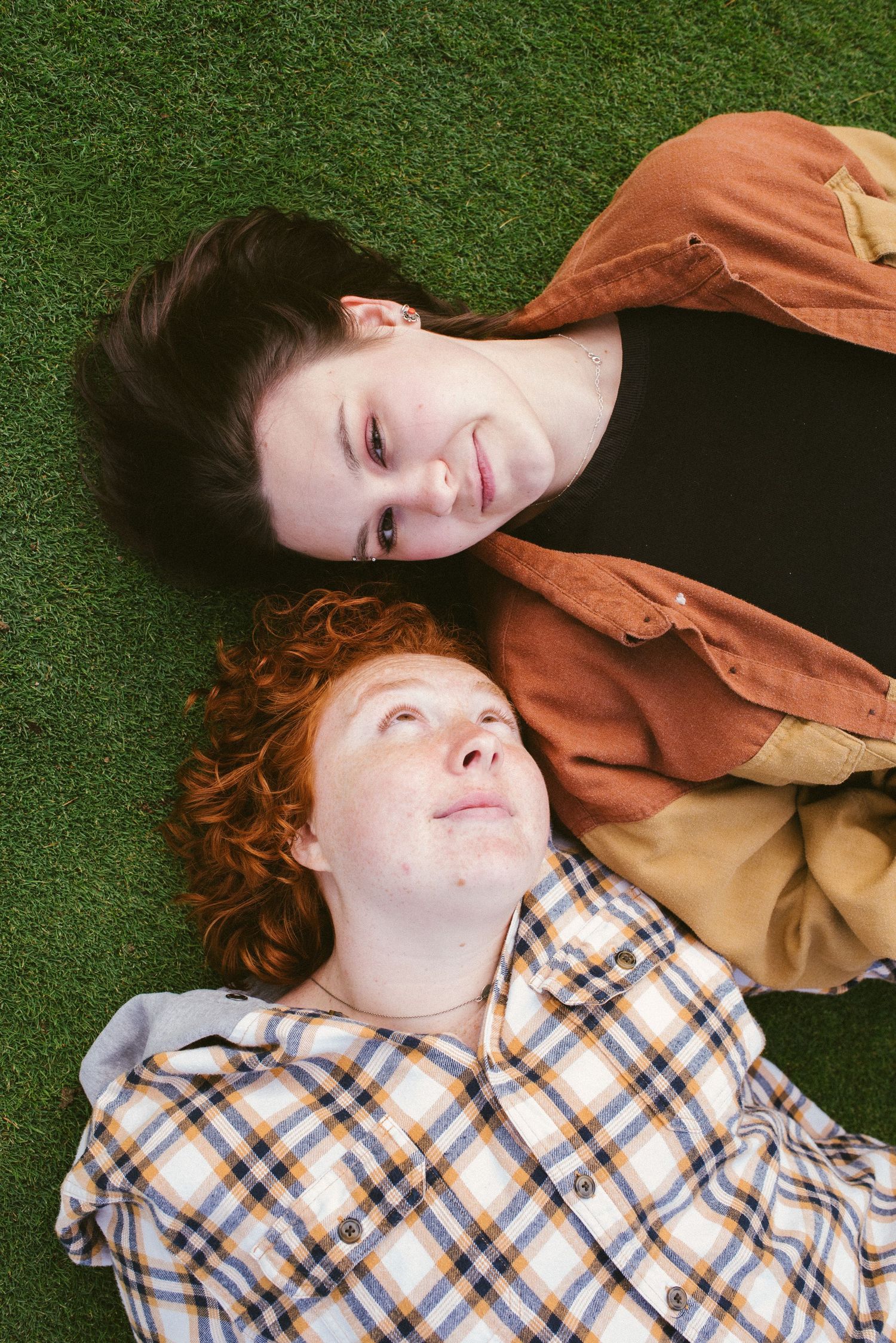 Two young people with red and dark hair lying on green grass, smiling and looking up in casual plaid shirts.