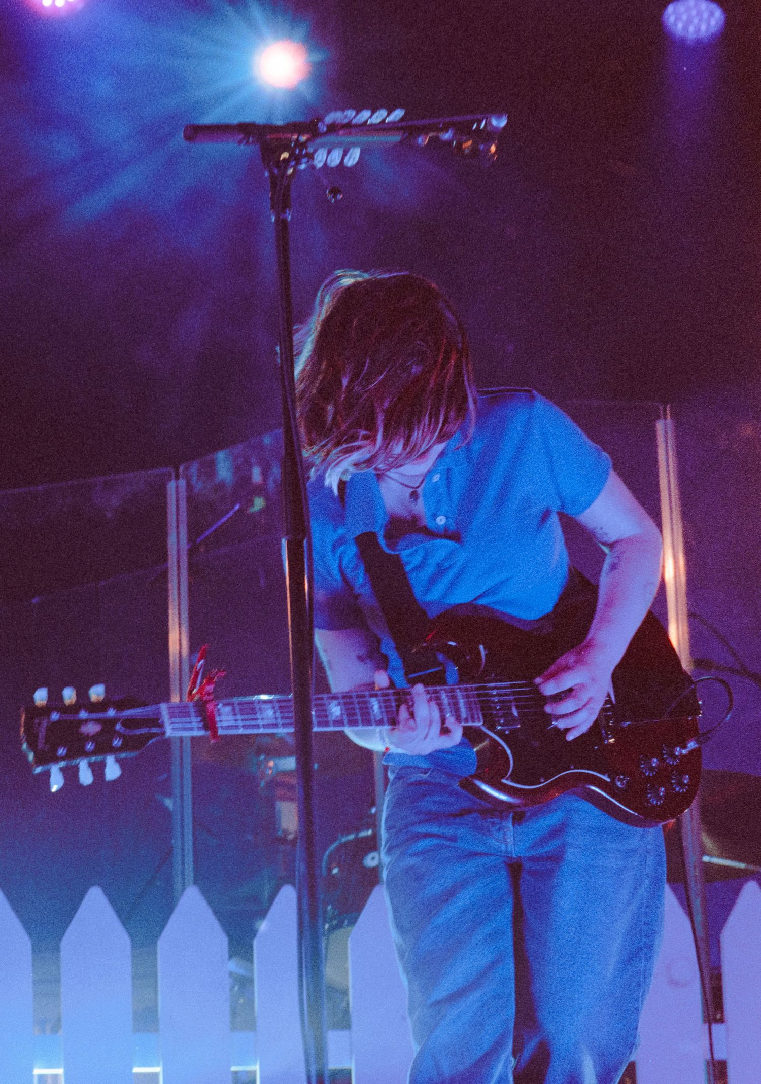 A guitarist with long hair performs on stage under blue lights, playing a black electric guitar while wearing a blue t-shirt.