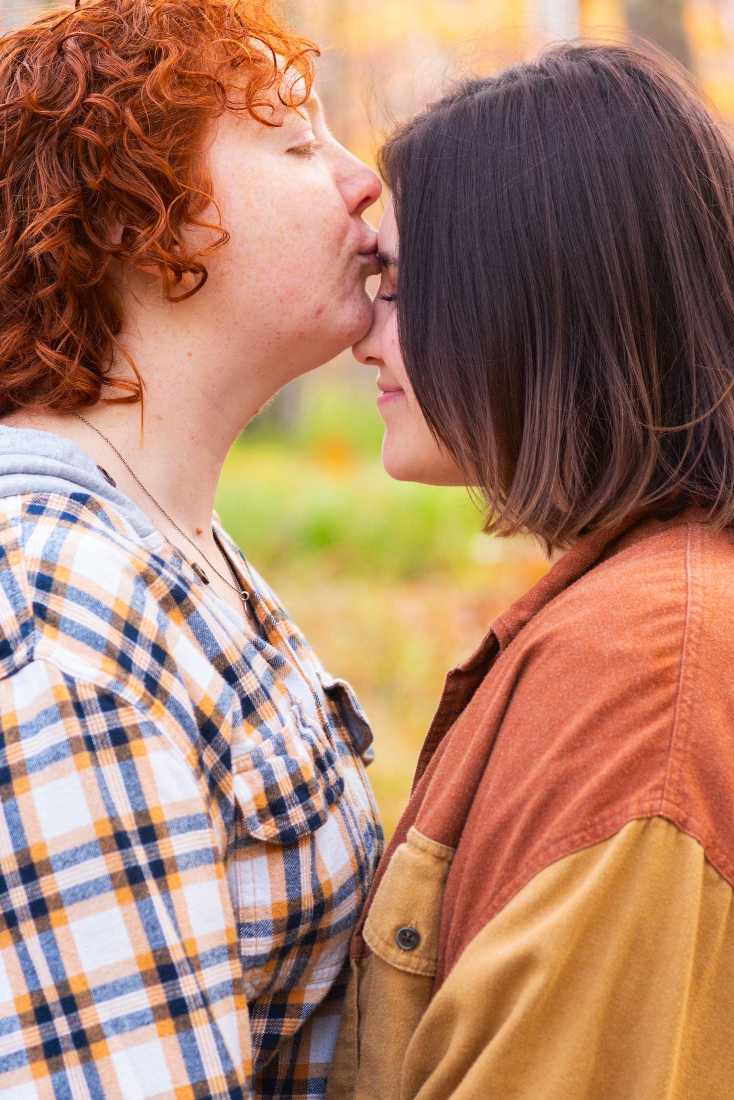 Two women sharing a tender kiss outdoors in autumn, one with curly red hair and one with dark hair.