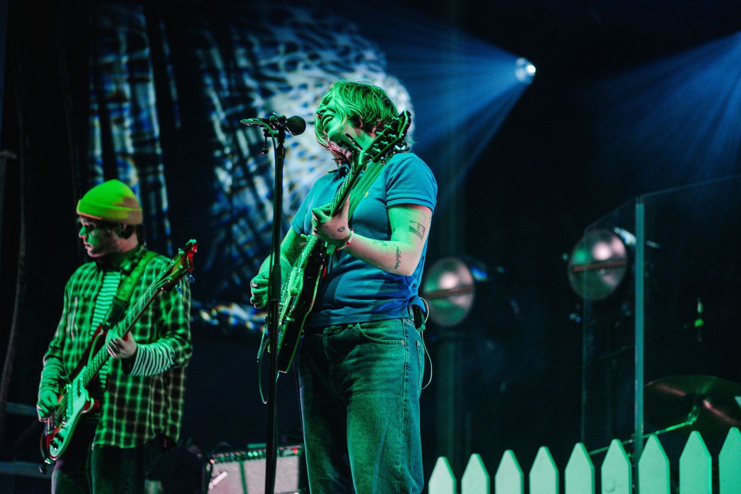 Two musicians performing on a green-lit stage with a white picket fence, playing guitars at a live concert.