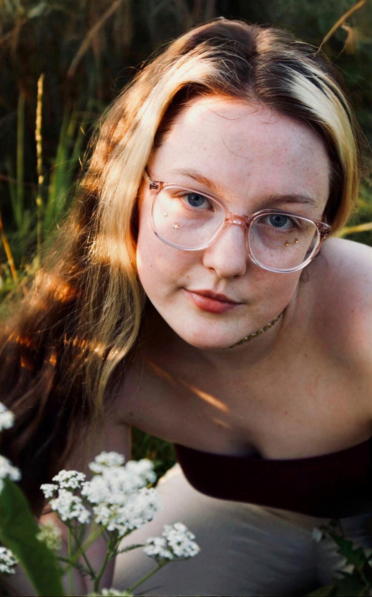 Young woman with pink glasses and freckles posing outdoors among green grass and white wildflowers.