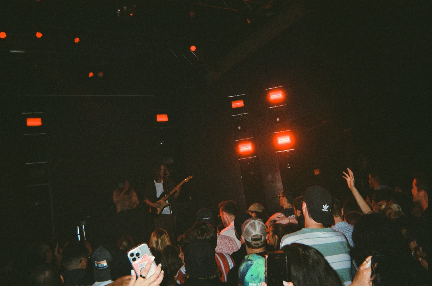 Dense concert crowd surrounds guitarist on stage under red overhead lights in a dark music venue.