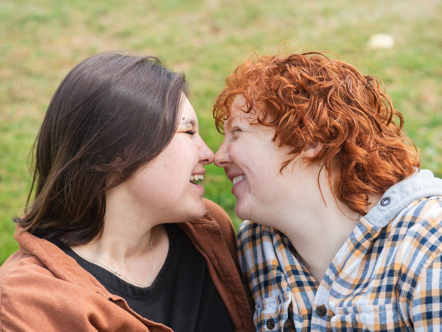 Two women smiling and touching noses affectionately outdoors, one with dark hair and one with curly red hair.
