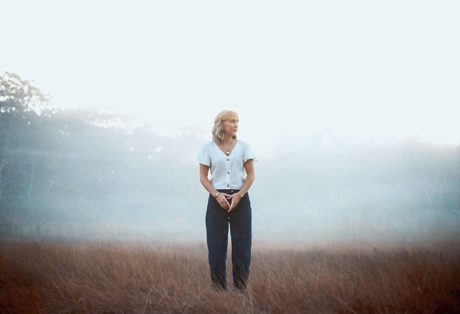 Blonde woman standing alone in misty foggy field wearing white shirt and dark pants.