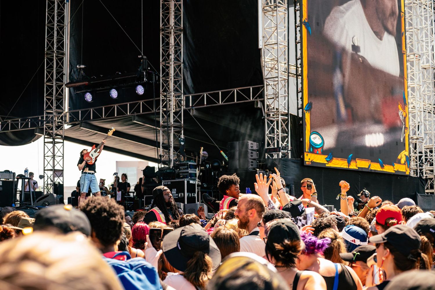 Performer on outdoor festival stage engaging with large crowd under bright sunlight.