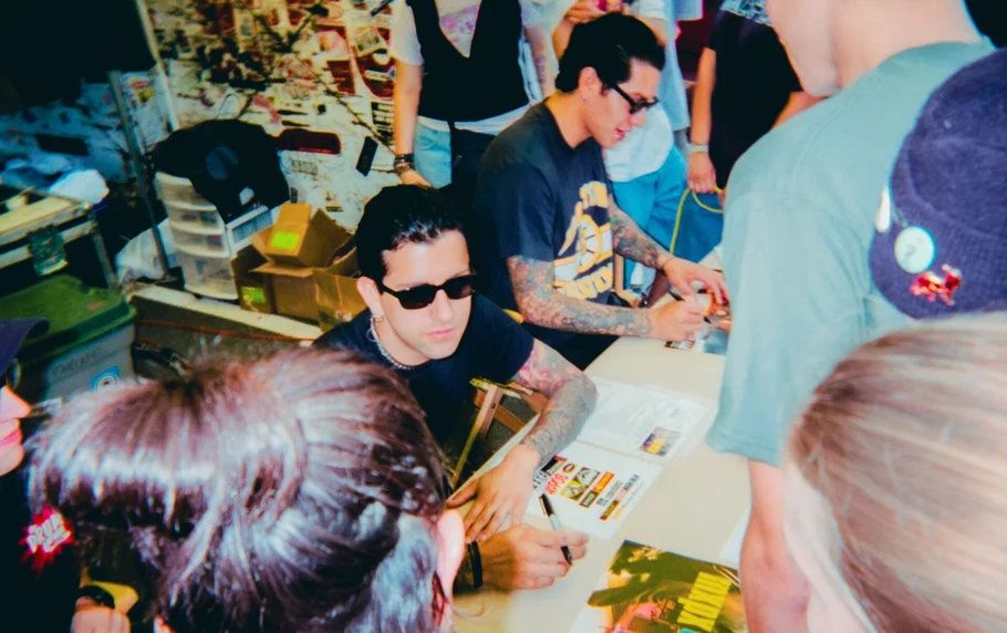 Musician in sunglasses signing autographs for fans at a merch table at Warped Tour festival.