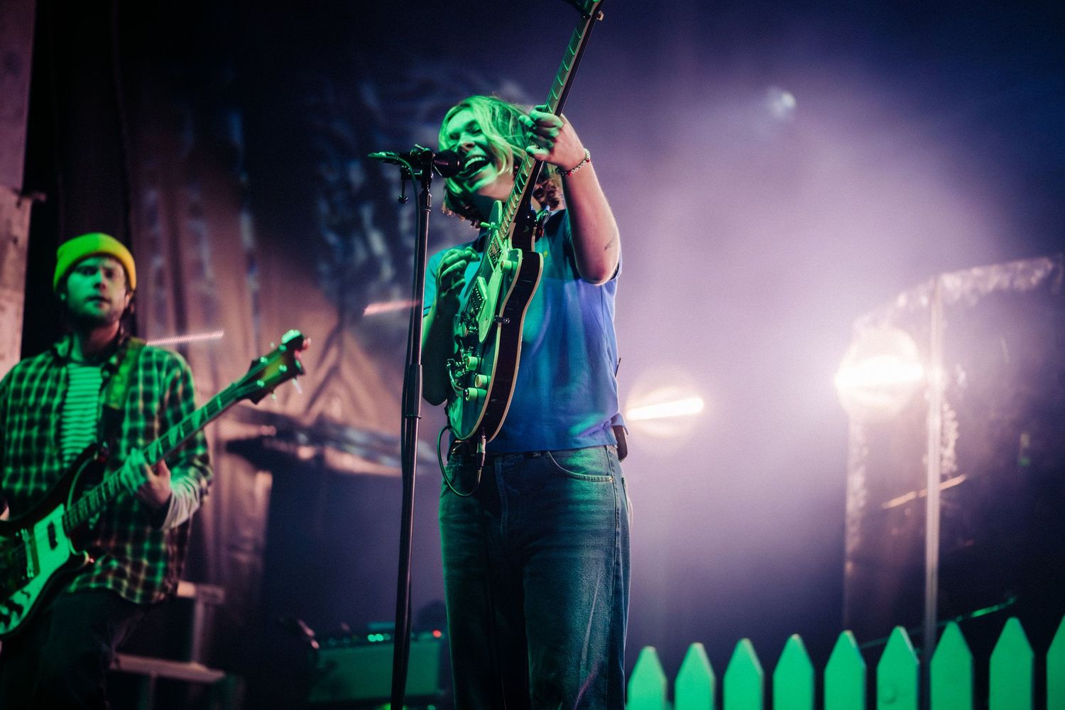 Two musicians performing on stage under green concert lights with foggy atmosphere.