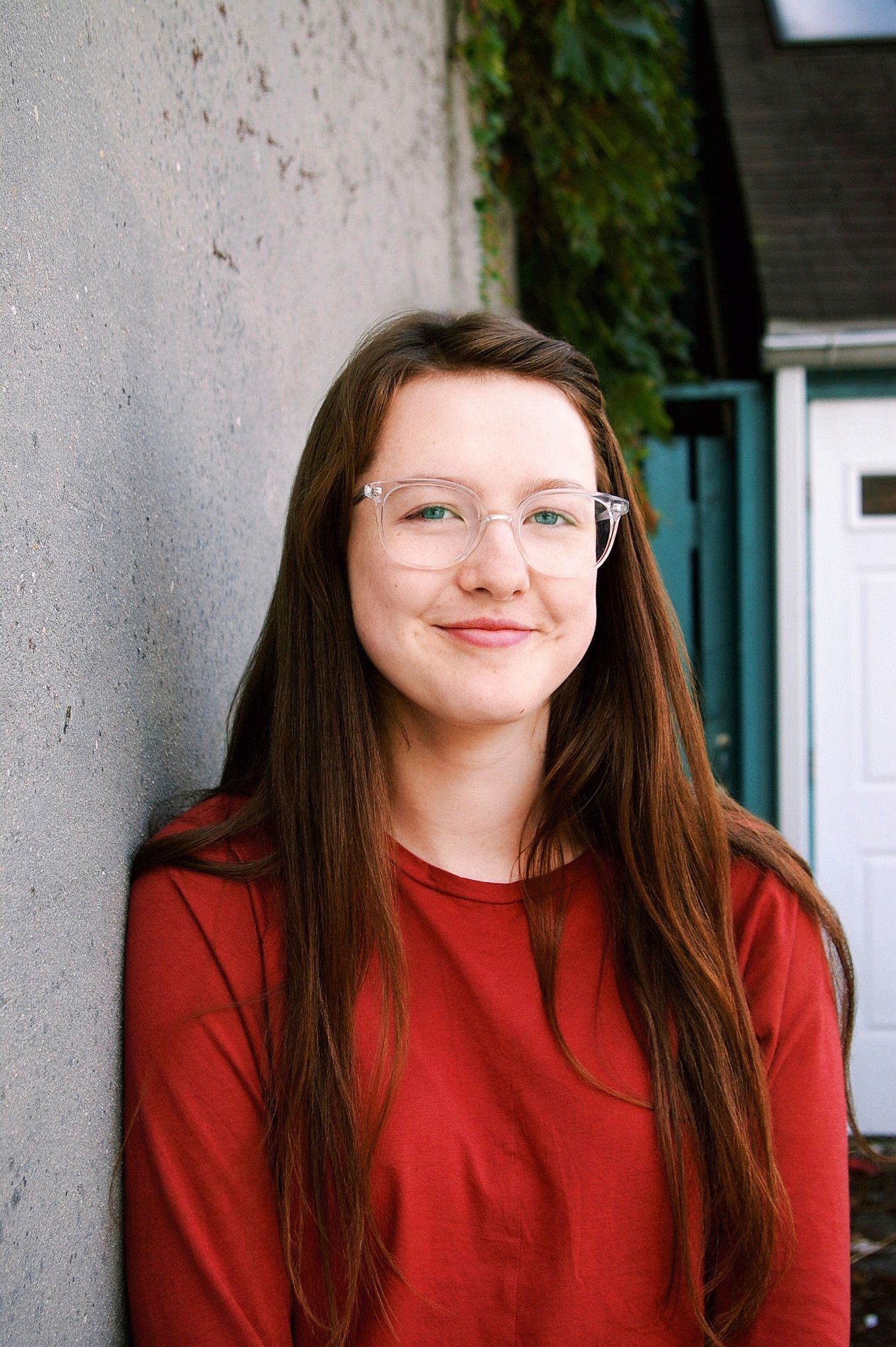 Young woman with long brown hair and clear glasses smiling, wearing a red shirt, leaning against a gray wall outdoors.
