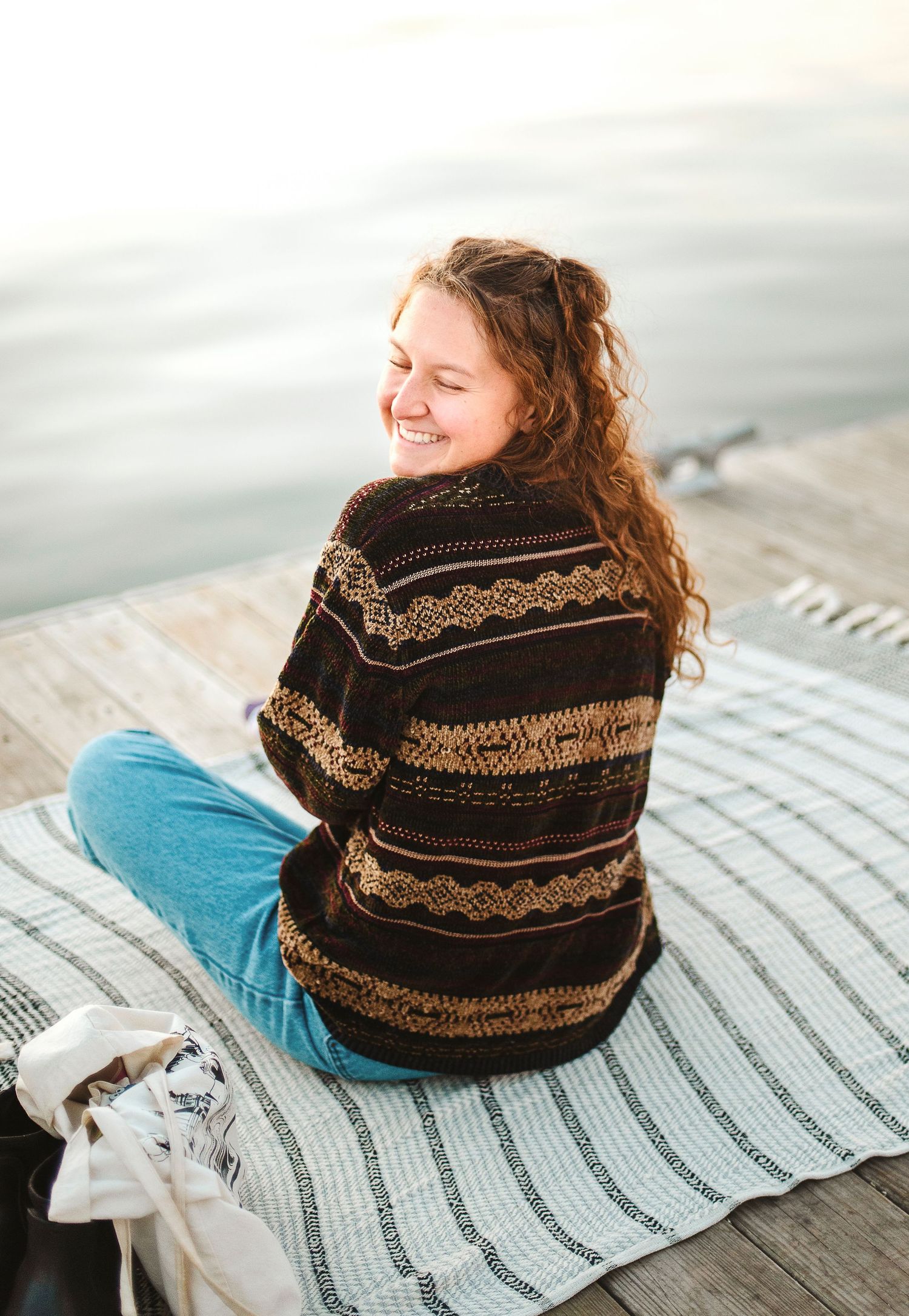 Smiling young woman with curly hair sitting cross-legged on a blanket on a wooden dock by the water.