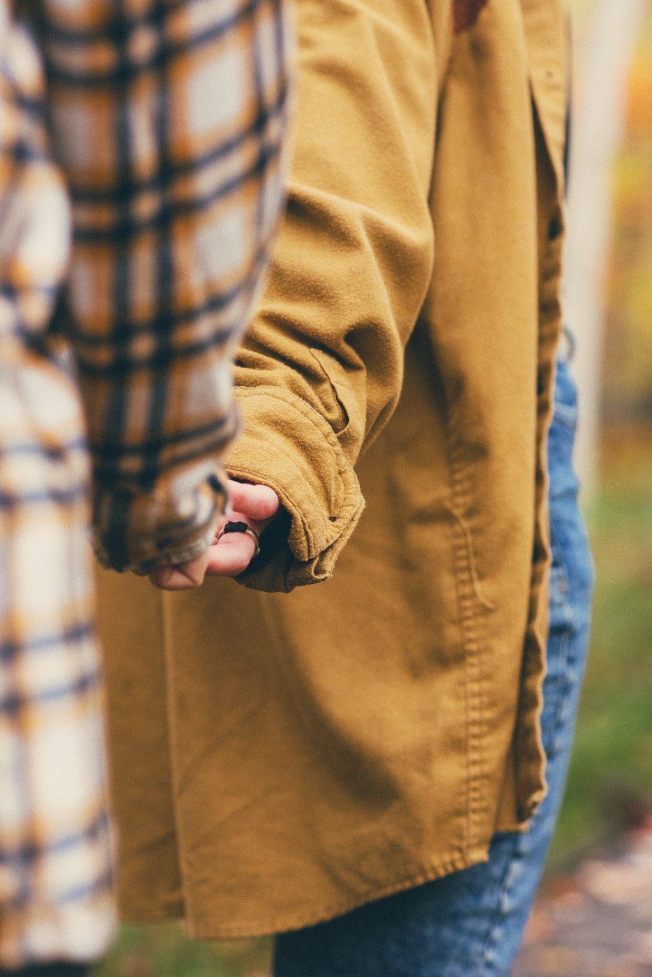 Two people holding hands outdoors, one wearing a tan jacket and jeans, the other in a plaid shirt, in an autumn setting.