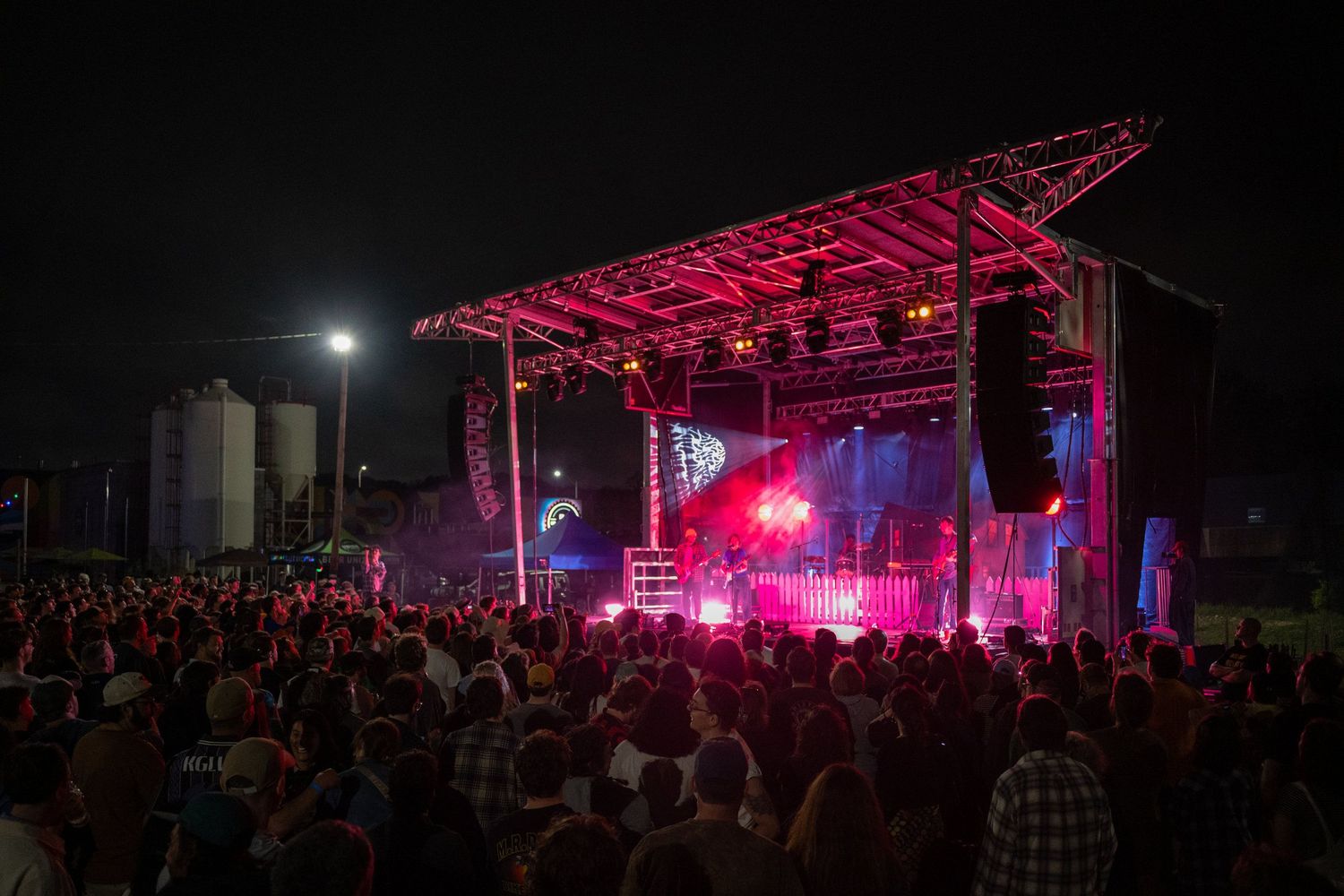 Outdoor concert stage lit with pink lights at night, packed crowd enjoying a live music festival performance.