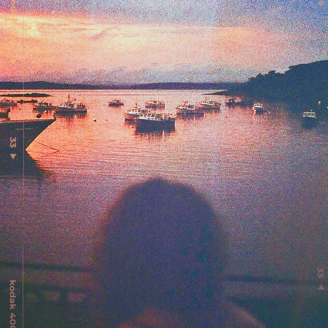 Silhouette of a person watching boats at sunset over a calm harbor with warm pink and orange hues on Kodak film.