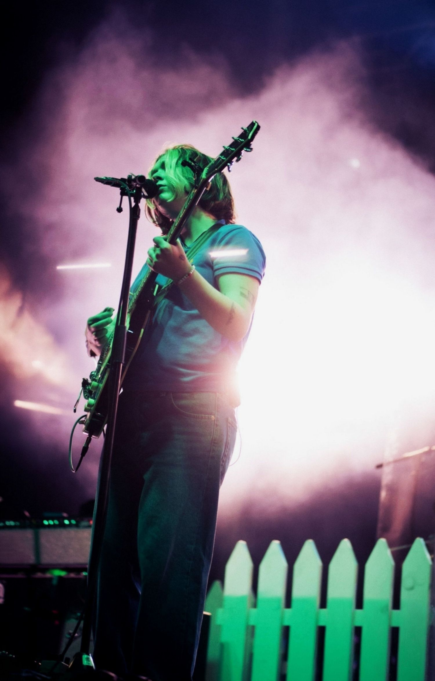 Guitarist singing into microphone on stage surrounded by dramatic purple smoke and bright lights.