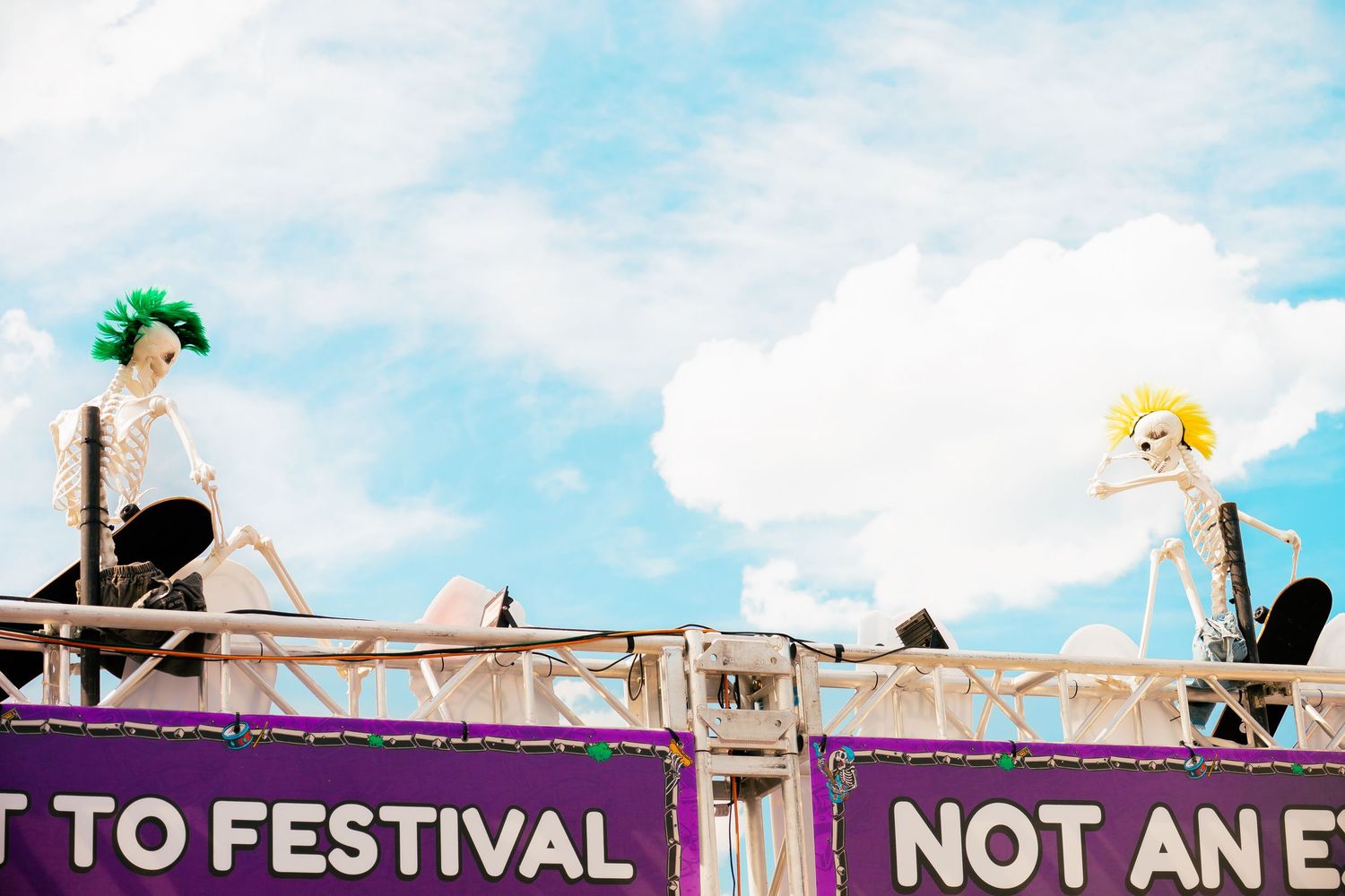 Two colorful skeleton decorations with mohawks displayed atop a purple festival sign under a cloudy blue sky.