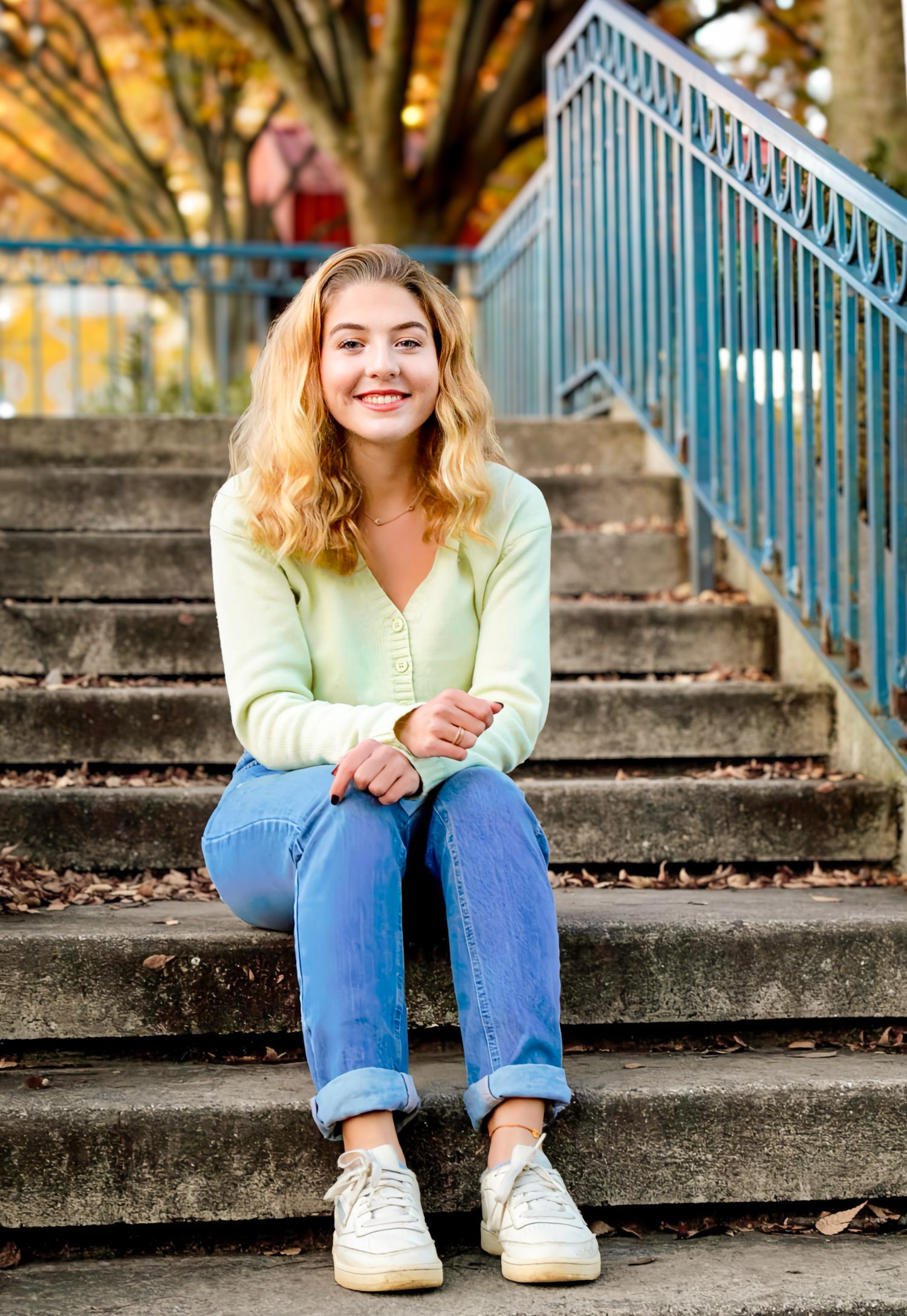 Young woman with curly blonde hair sitting on outdoor steps, wearing a mint green cardigan and jeans, smiling.