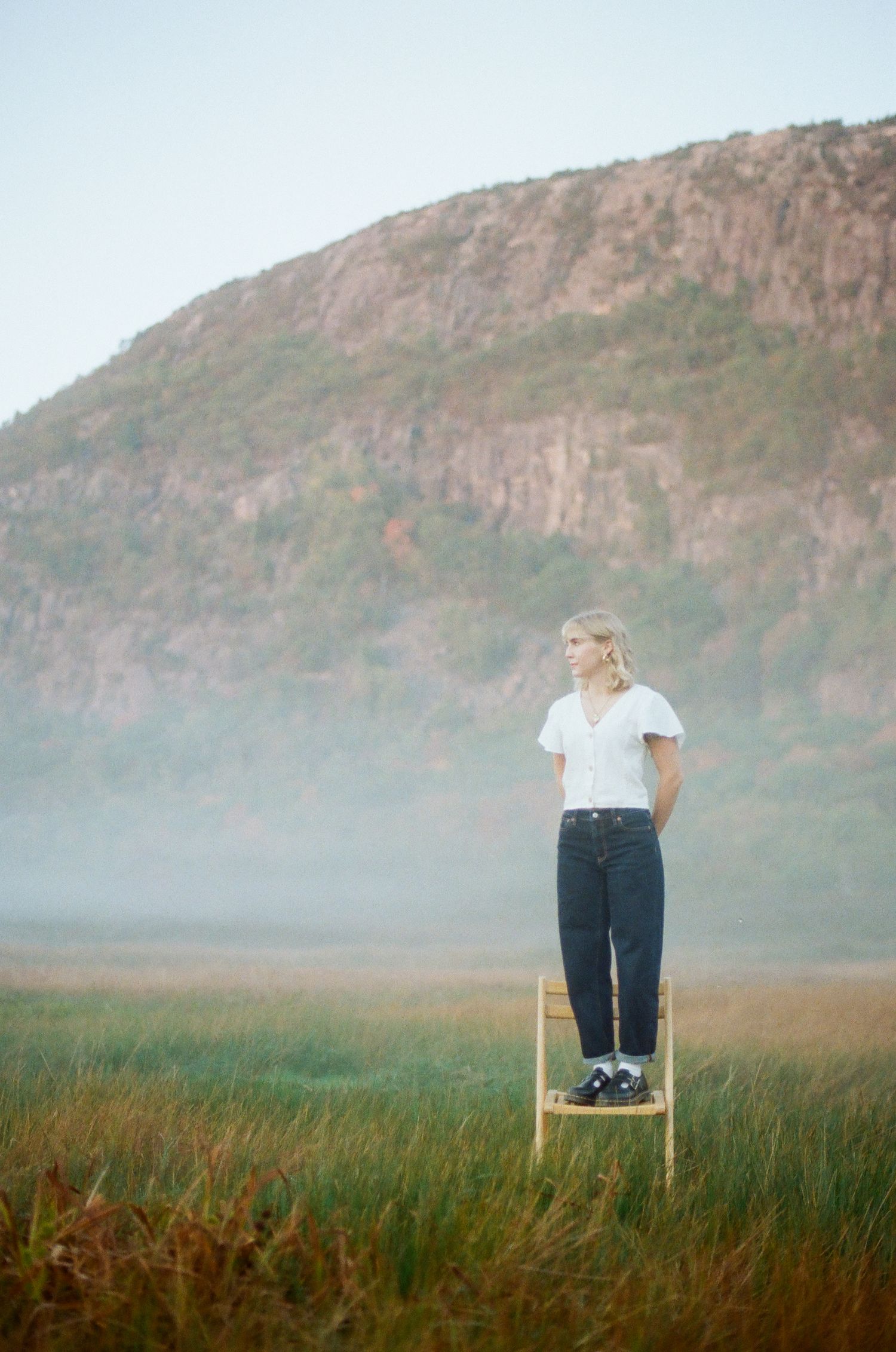 Woman in white top and jeans standing on a ladder in a misty field with a rocky hill backdrop.