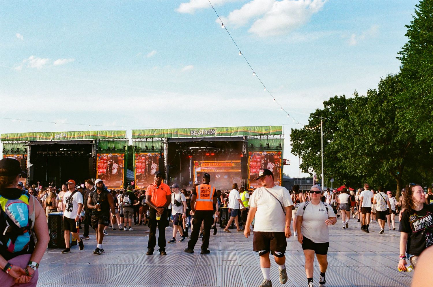 Crowds gather at an outdoor music festival with a large stage, green banners, and security staff in orange vests.