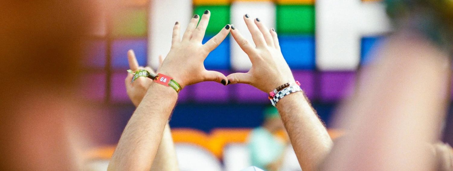 Close-up of raised hands with colorful festival wristbands and painted nails against a rainbow backdrop.