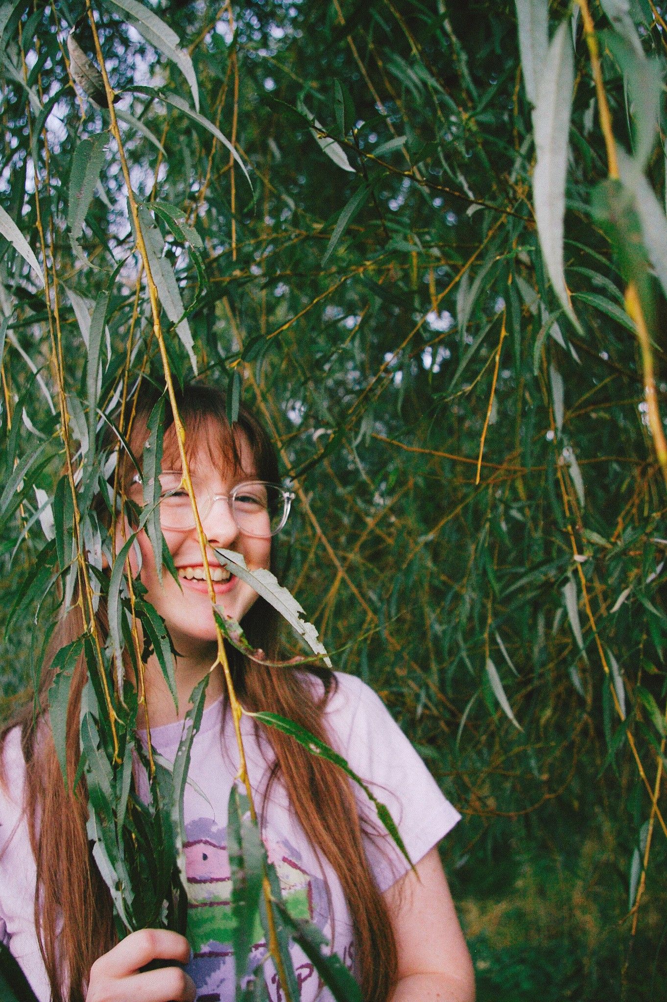 Smiling girl with long hair peeking through green willow tree branches outdoors.