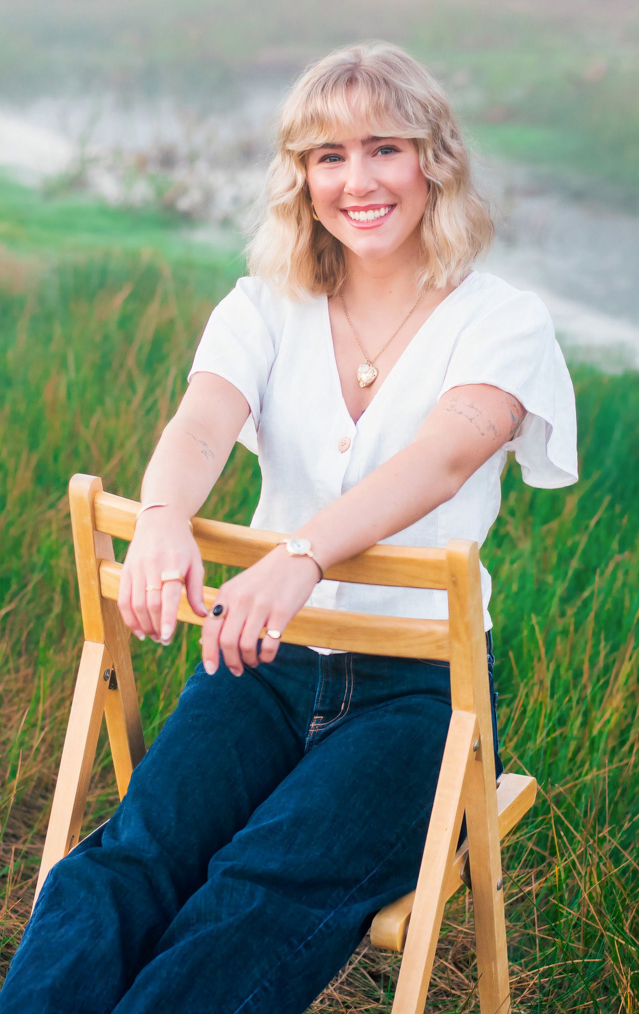 Smiling young blonde woman in white top and jeans sitting on wooden chair in green grassy outdoor setting.