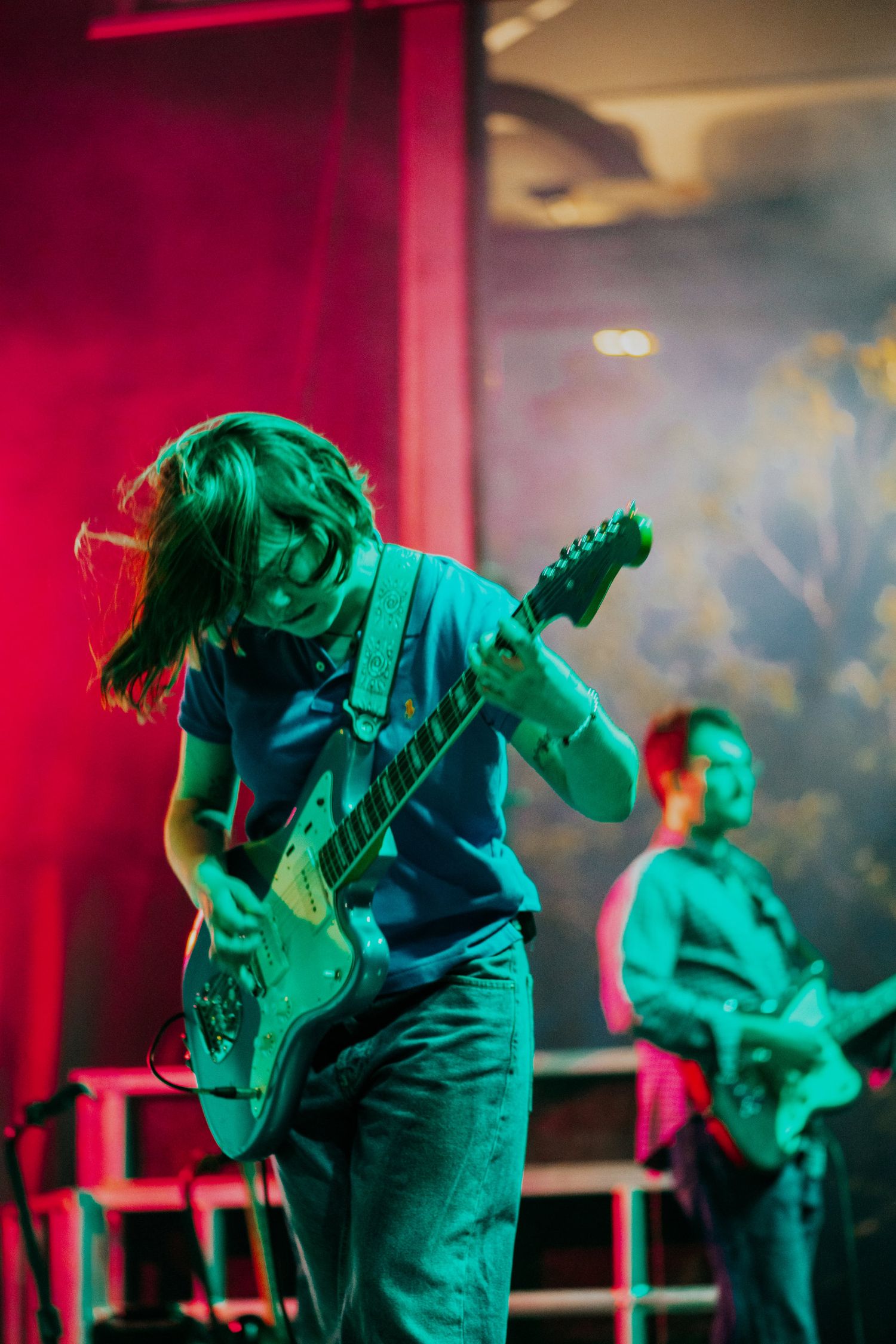 Two guitarists performing on stage under dramatic red and green lighting with atmospheric smoke effects.