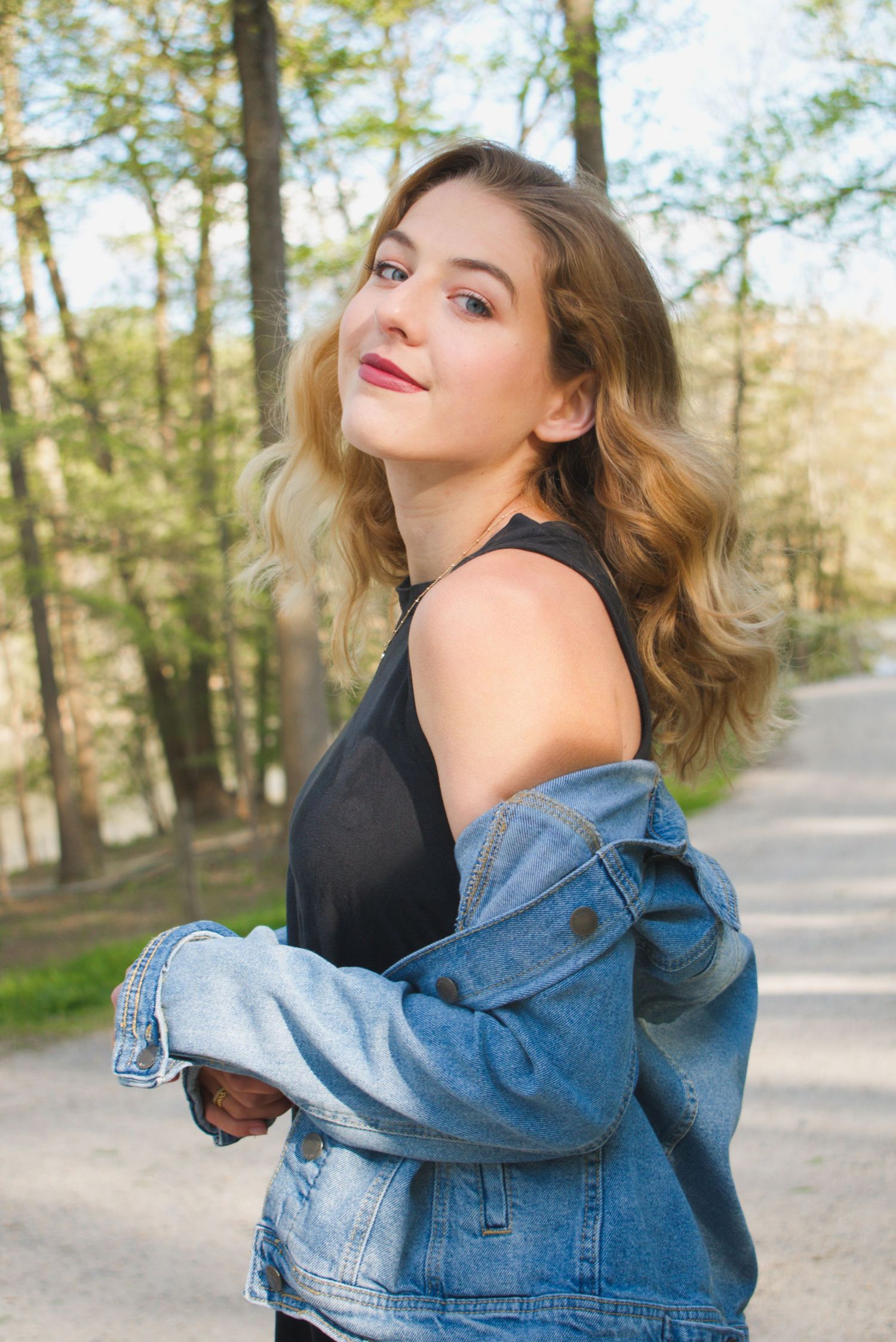 Young woman in denim jacket and black top smiling outdoors in a sunlit forest setting.