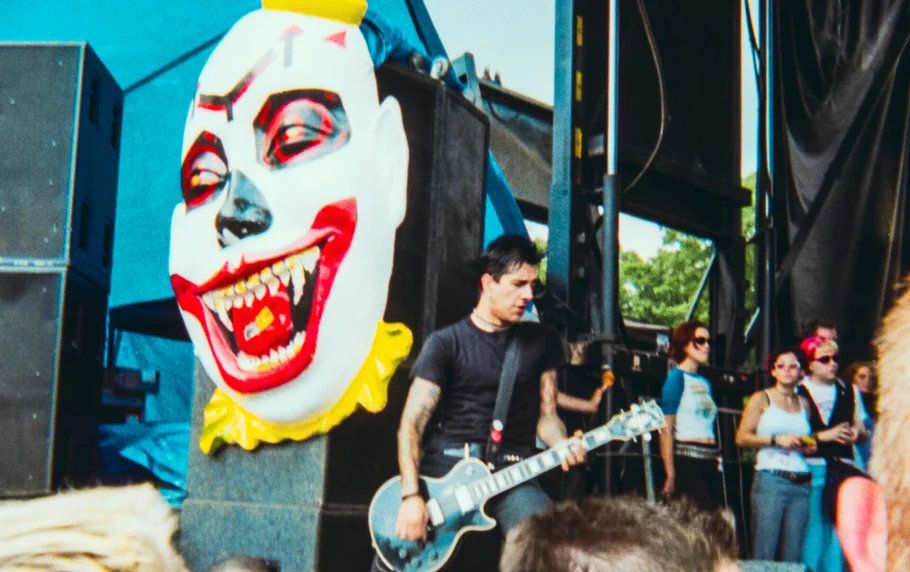 Guitarist performs on stage in front of a giant clown face backdrop at an outdoor music festival.